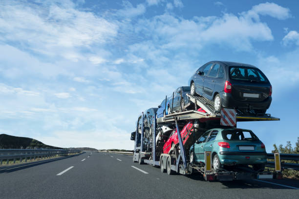 A car carrier truck, part of NY Auto Transport Fulton, transports several cars on an open highway under a blue sky with scattered clouds. The road is mostly empty, with safety barriers on the sides.