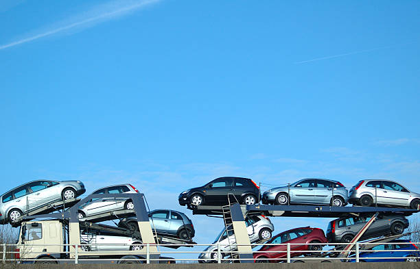 A car carrier truck loaded with multiple cars is driving on a road under a clear blue sky. The cars, stacked in two levels on the trailer, illustrate the efficient services provided by NY Auto Transport Fulton for dependable car transport in the region.