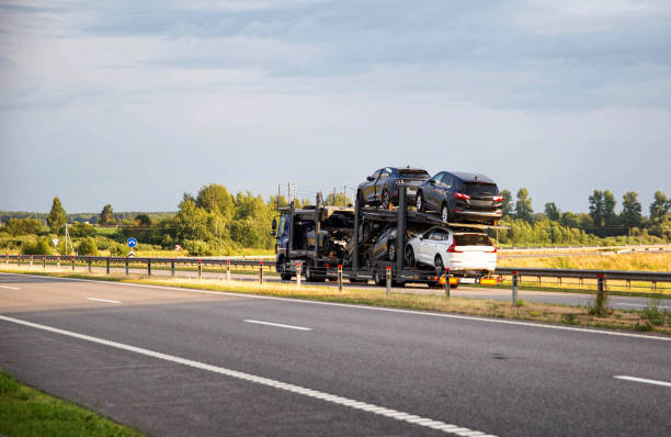 A car carrier truck loaded with multiple vehicles drives on a highway surrounded by greenery and trees under a partly cloudy sky, showcasing reliable Car Transport Fulton, NY services in action.