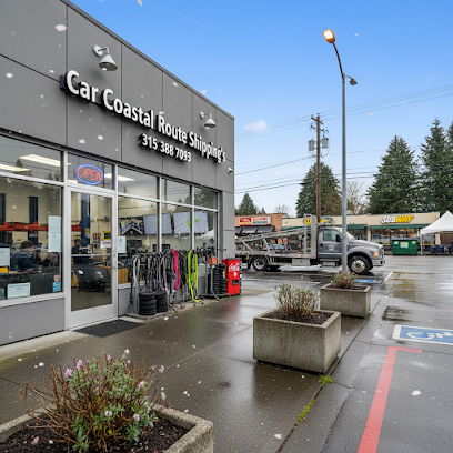 Car Shipping Fulton, NY storefront on a wet day with a tow truck parked outside, plants in concrete planters, and nearby stores, including Subway, visible across the street. Light snow is falling, adding to the cozy atmosphere of this Car Transport Fulton, NY service location.
